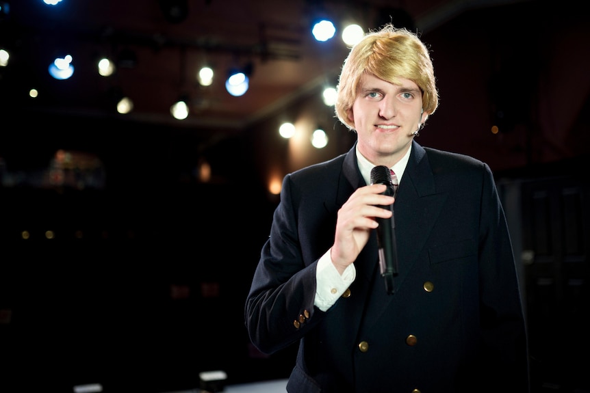 A teenage boy dressed in a blonde bowl cut wig and suit holding a microphone, smiling at the camera