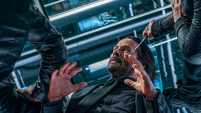 Colour still of Keanu Reeves held down on the floor, arms raised, two men stand over him with a knife in John Wick: Chapter 3.