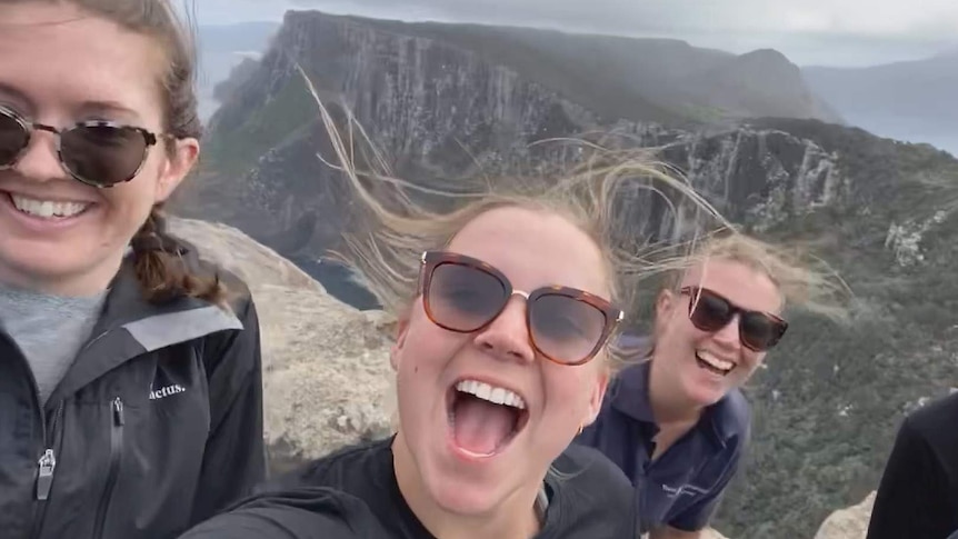 Three young women smiling wildly in the wind above a rugged coastline, a sheer cliffed island offshore