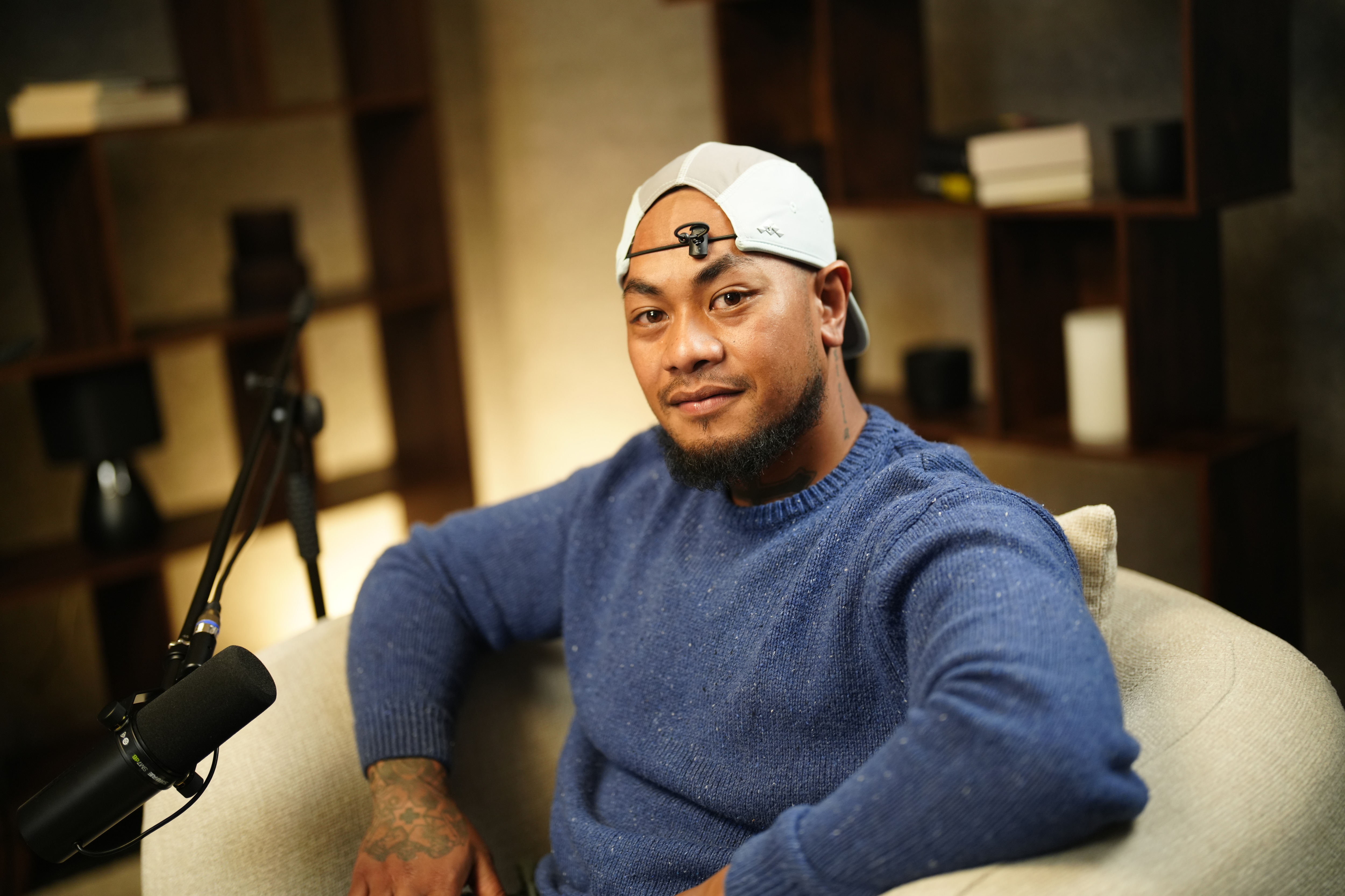 A man sitting in an armchair wearing a blue jumper and a backwards white cap.