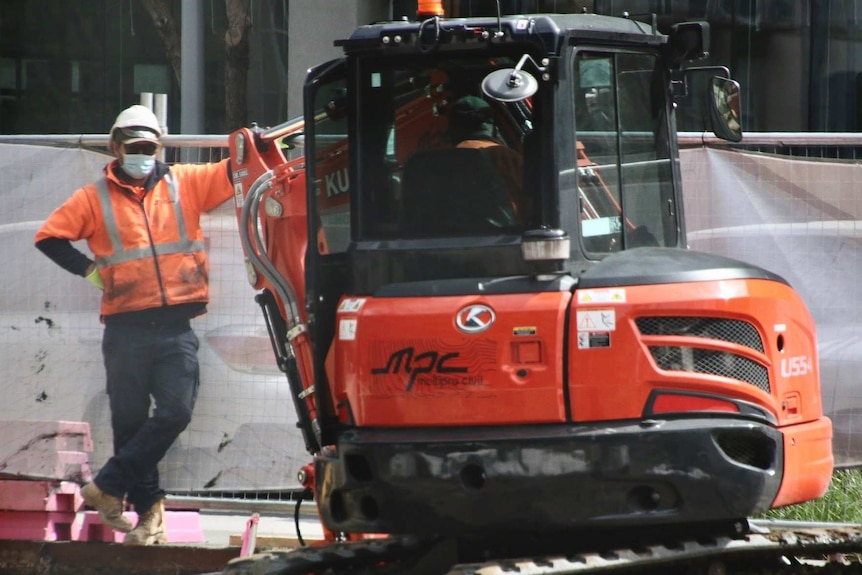 A construction worker wearing a mask, helmet and orange fluro top leans against a small excavator.