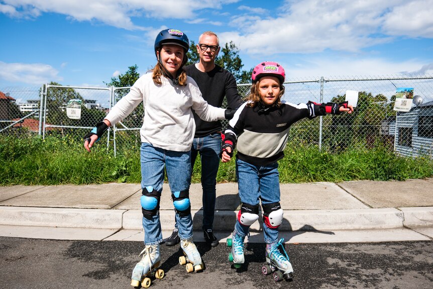 An older man in glasses stands behind his two children who are wearing roller skates and helmets