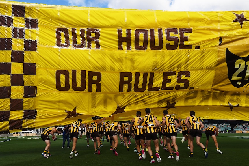 Hawthorn players run through a banner that reads 'Our House Our Rules' in an AFL game in Tasmania.