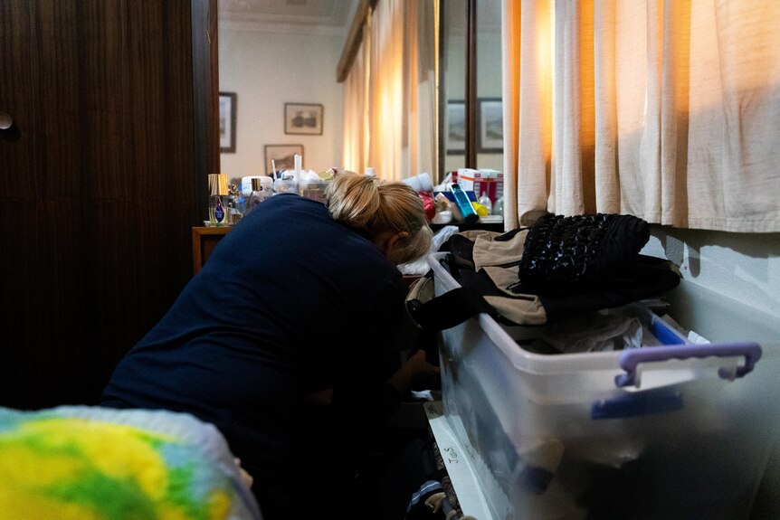 A woman crouched down in front of a cluttered vanity table, sifting through things.