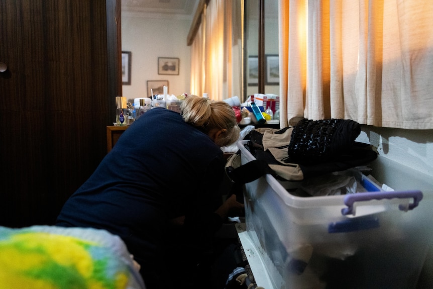 A woman crouched down in front of a cluttered vanity table, sifting through things.