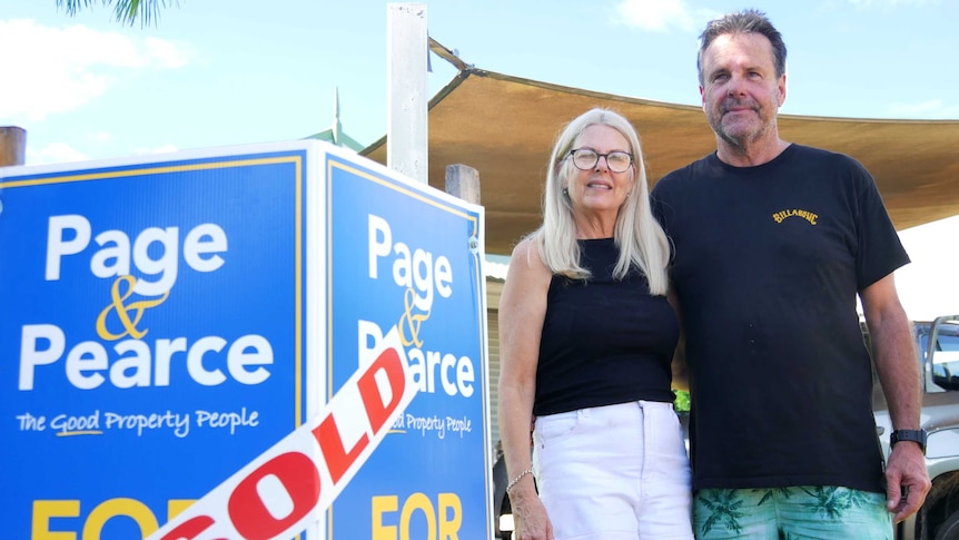 A man and woman stand with their arms around each other next to a 'sold' sign outside a house.