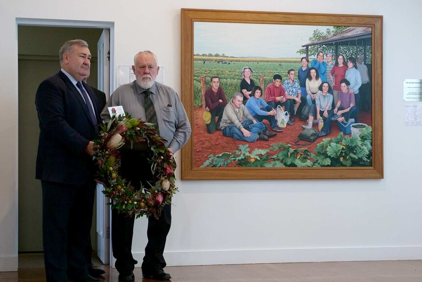 Two men stand holding a wreath with a large painting of farm workers.