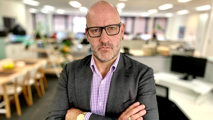 John Winter stands with arms folded in an office with desks and computers in the background.