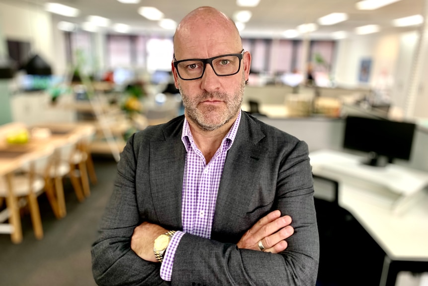 John Winter stands with arms folded in an office with desks and computers in the background.