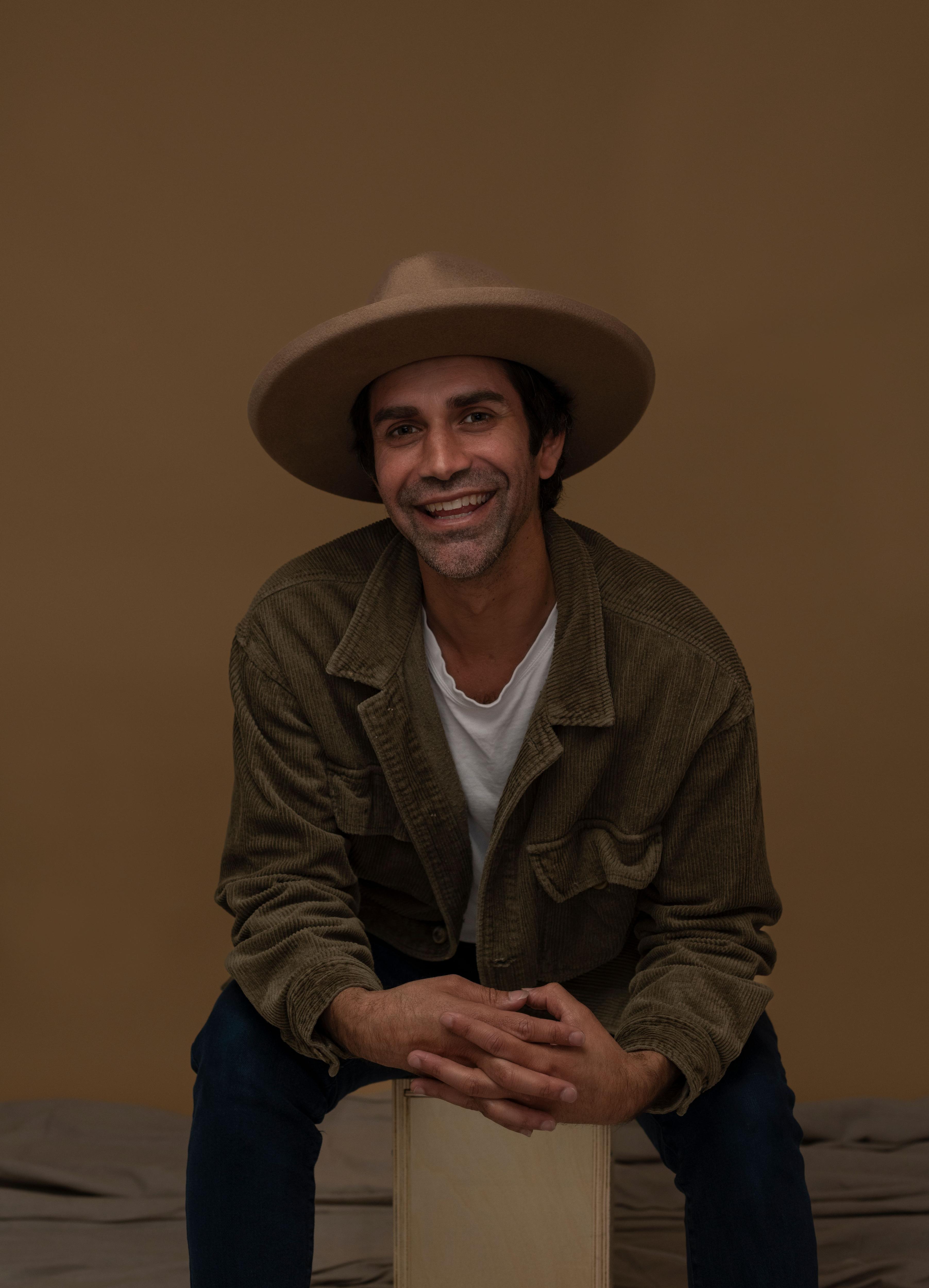 Pirritu is smiling at the camera. He is wearing a wide brim beige hat and a khaki shirt. 