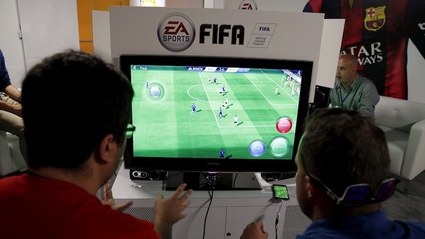 Two men sit in front of a video game console with EA and FIFA branding.