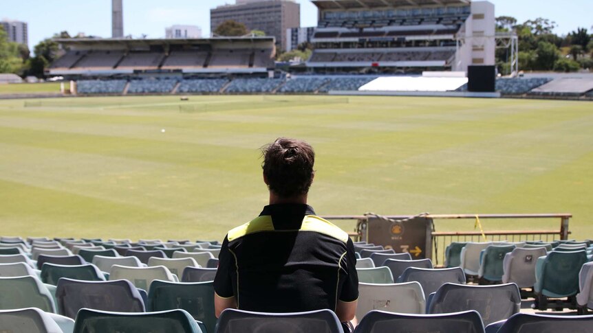 A wide shot of Hilton Cartwright sitting in the stands at the WACA Ground with his back to the camera looking out over the oval.