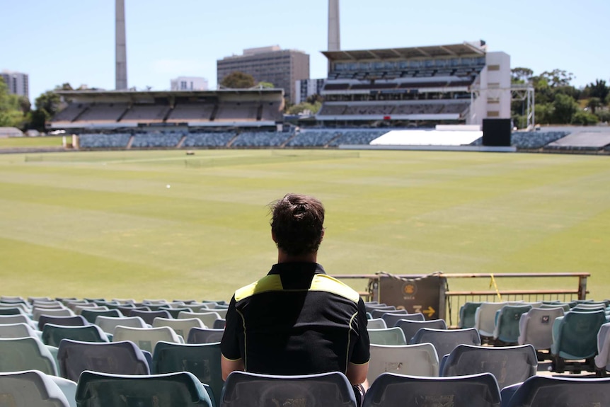 A wide shot of Hilton Cartwright sitting in the stands at the WACA Ground with his back to the camera looking out over the oval.
