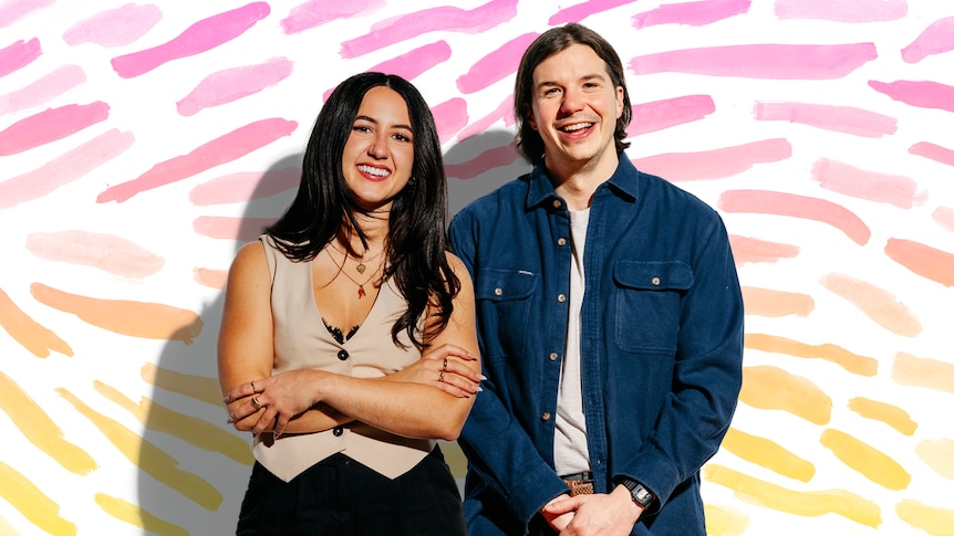 Two people stand in front of a white background with watercolour brushstrokes on it 