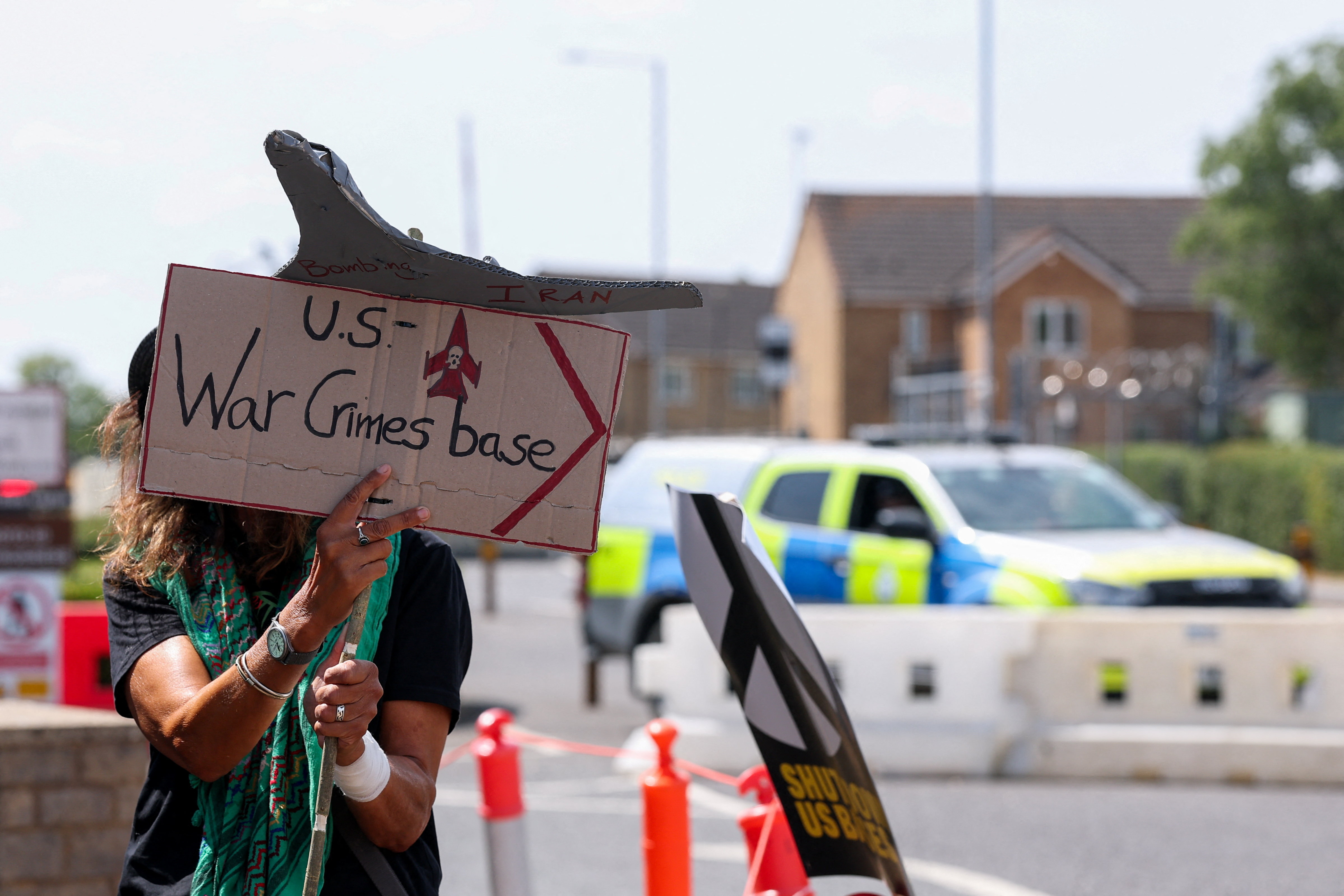 A person holds up a cardboard protest placard saying "US War Crimes Base".