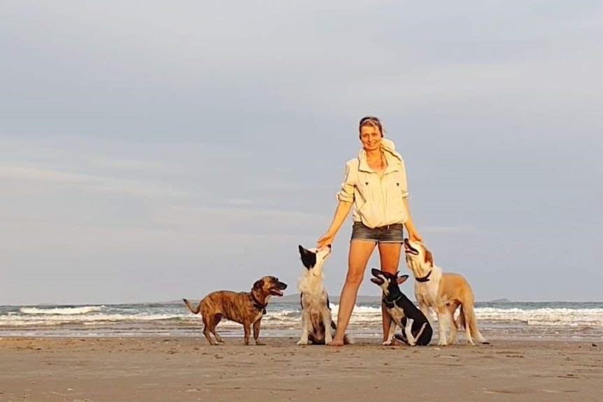 A woman stands on the beach with four dogs looking up at her.