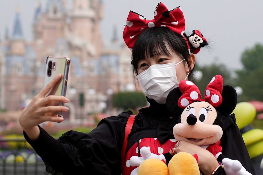 A woman with a plush Minnie toy takes a selfie with a Disney castle in the background