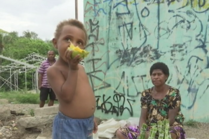 A small child eating a piece of fruit, with a woman sitting on the ground selling goods.