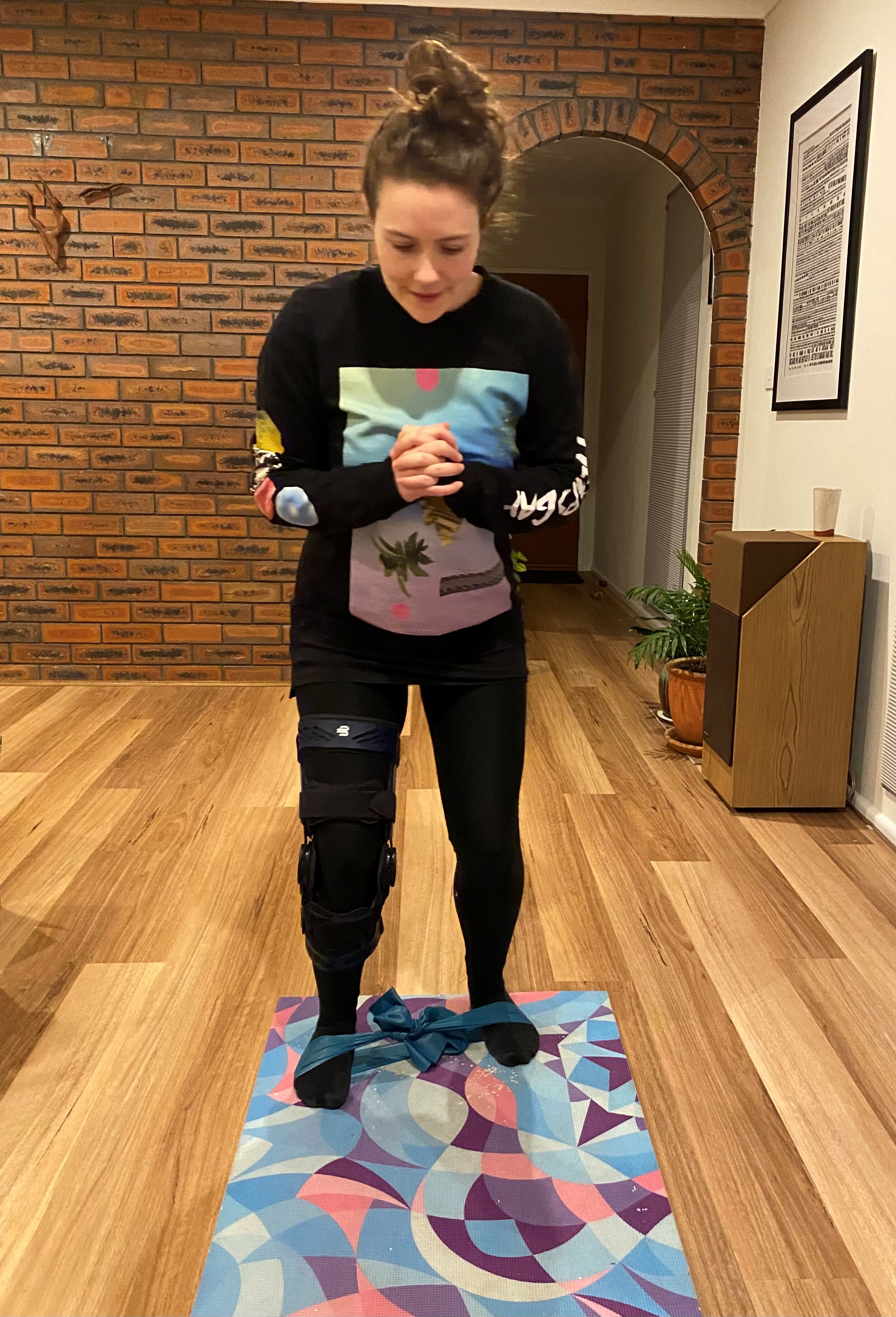 A woman with a knee brace on her right leg, her hands clasped together walks along a mat with a band keeping her feet together