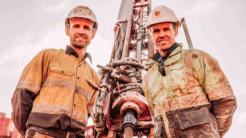 Two men wearing high-vis workwear standing in front of a large drill rig used in minerals exploration