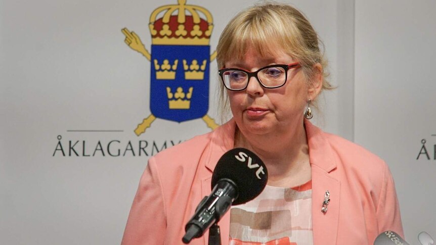 A woman wearing a blaze stands in front of background with Swedish crests, microphones aimed at her.