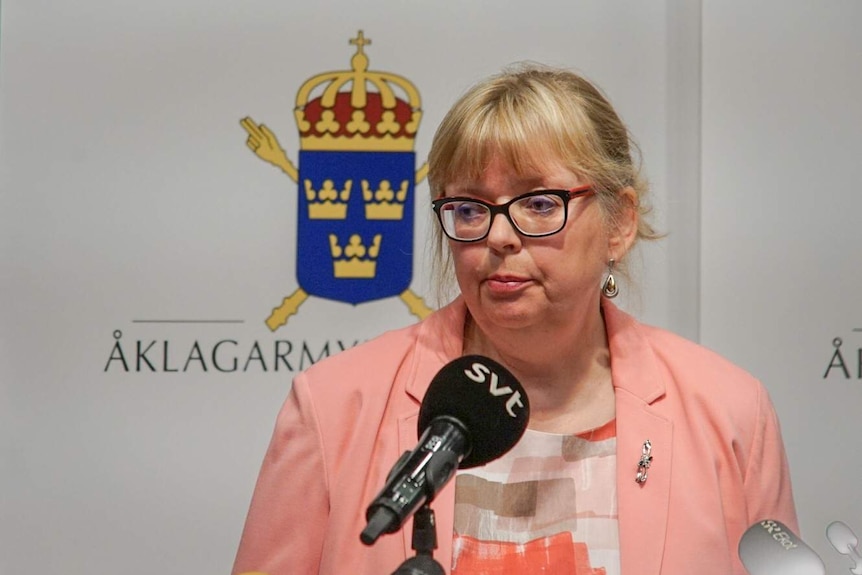 A woman wearing a blaze stands in front of background with Swedish crests, microphones aimed at her.