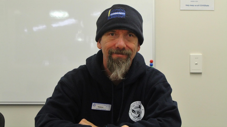 A man wearing  navy blue jumper and beany smiles at the camera. His beard has a mix of black and grey hair. 
