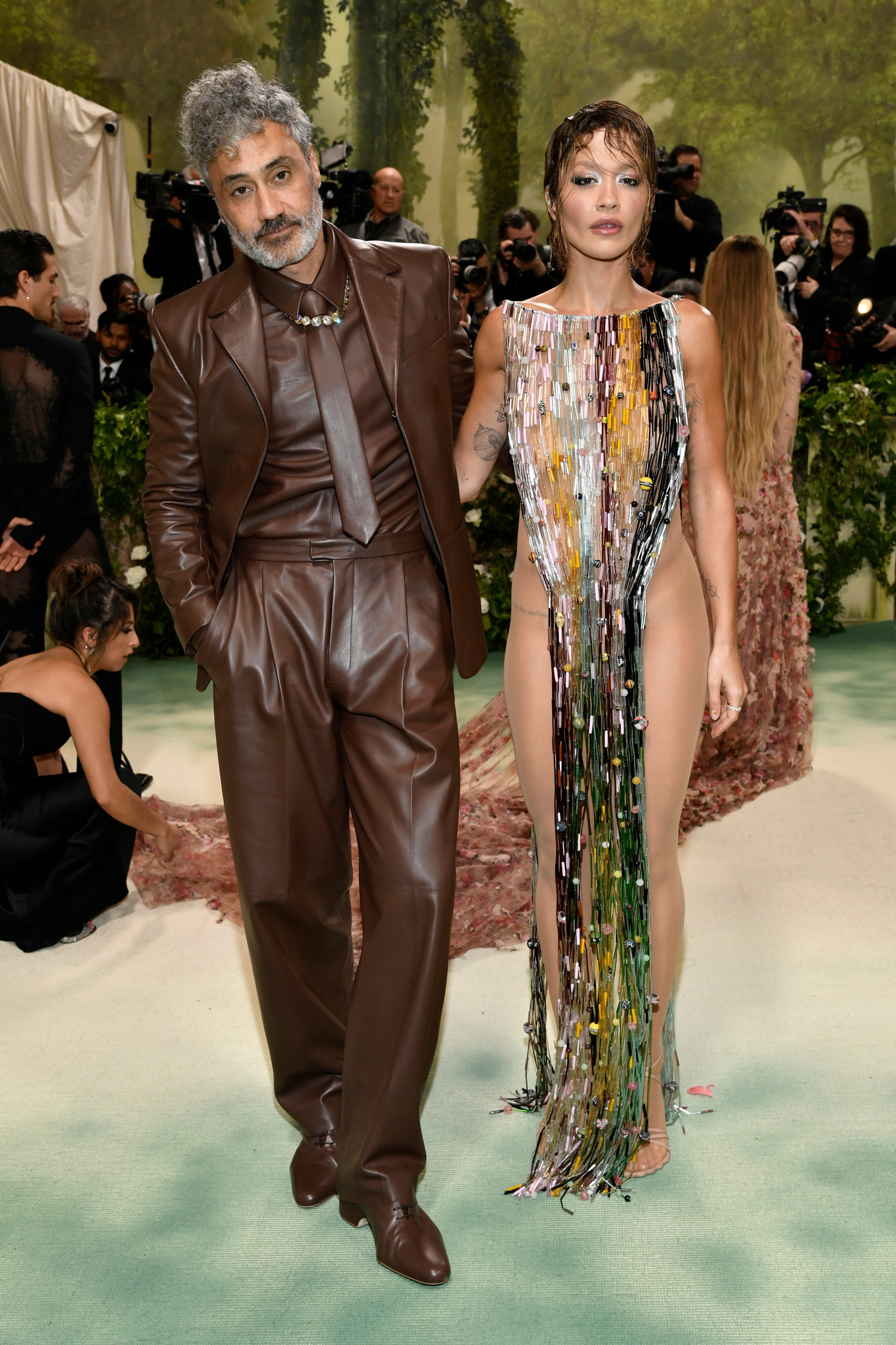 Taika Waititi wears a brown leather suit with a matching tie and Rita Ora in a dress made from a curtain of coloured beads