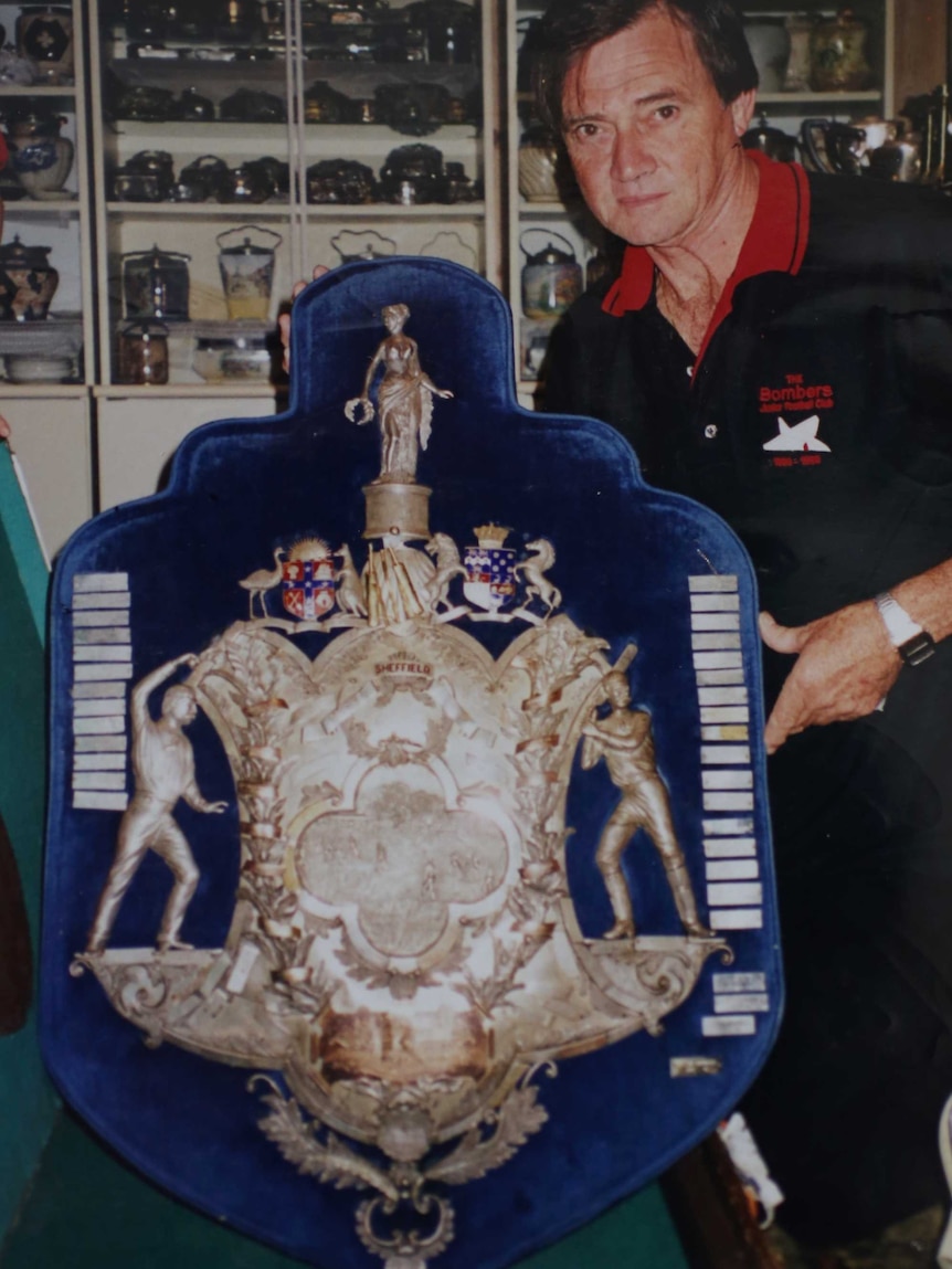 A man in a blue and red polo shirt stands holding the cricket Sheffield Shield.