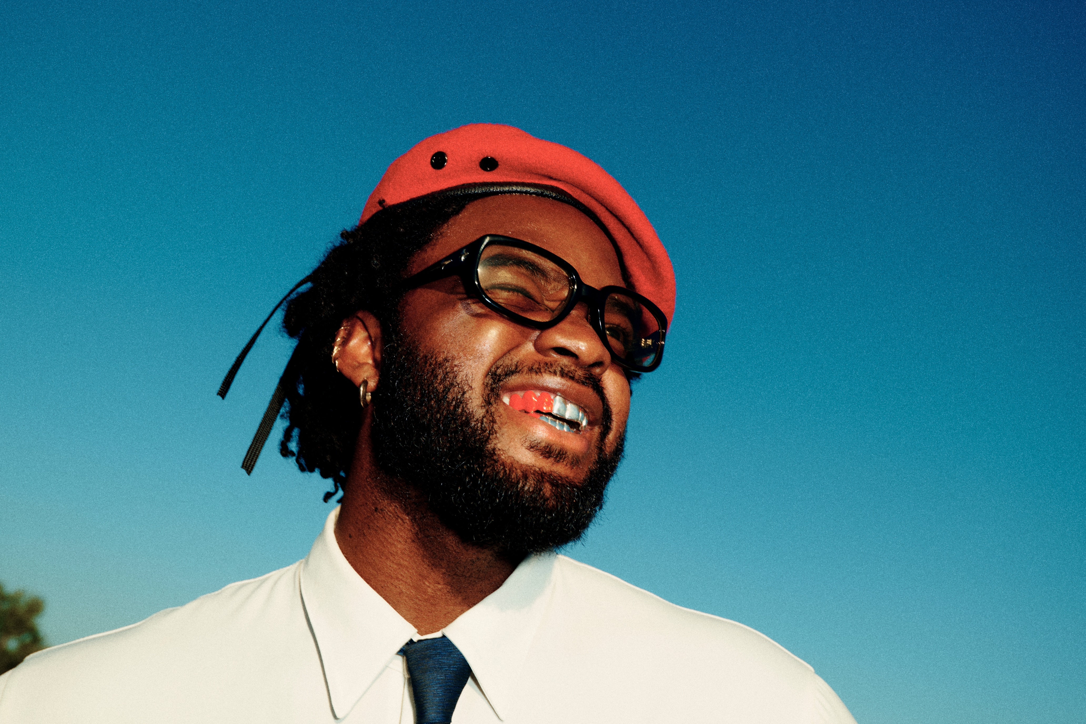 Genesis Owusu smiles and squints against a blue sky. He's wearing a red beret, thick rimmed glasses and red paint on his teeth