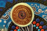 A coin, inset into an Indigenous artwork. Both the coin and the painting feature a handprint.