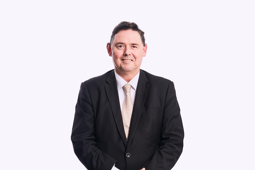 Portrait of Shane Kelly smiling and wearing suit in front of white background.