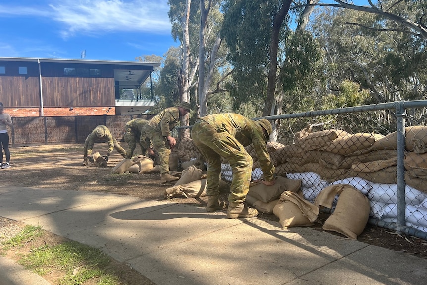 Army members in uniform shift sand bags along a fence.