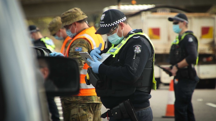 Men in navy blue police uniforms and khaki ADF gear wearing blue surgical masks checking cars at a roadblock.