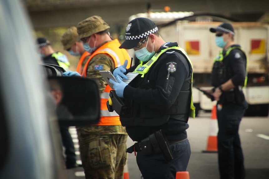 Men in navy blue police uniforms and khaki ADF gear wearing blue surgical masks checking cars at a roadblock.