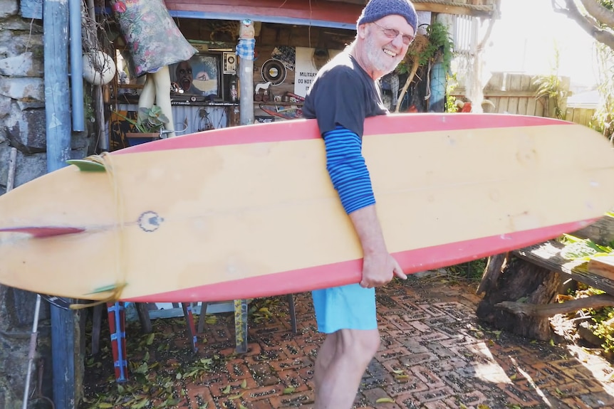 A man in boardshorts and beanie smiles, holding an old surfboard
