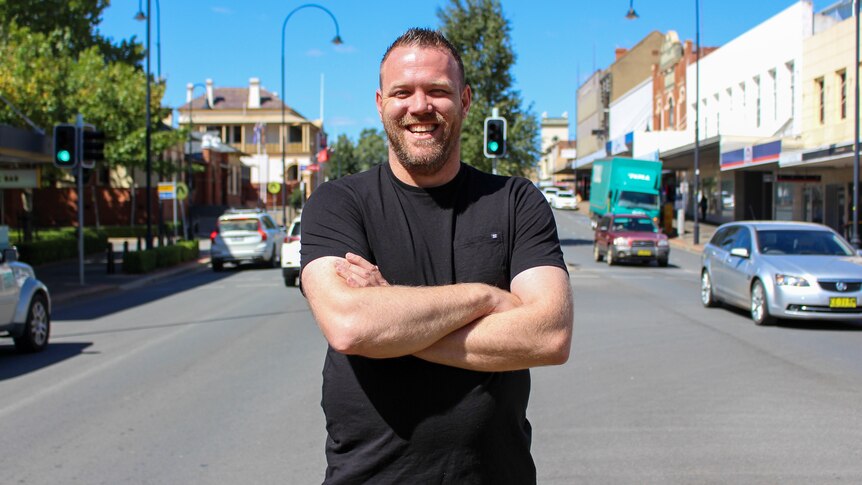 A man with short spiky hair and a beard, wearing a black t-shirt, standing with arms folded in the street of a country town.