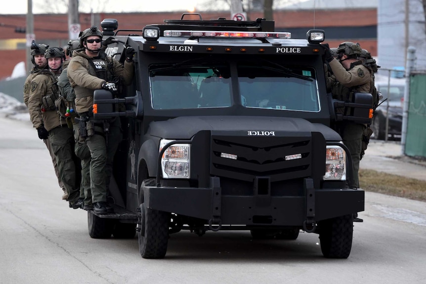 Up to eight police officers wearing khaki gear with dark helmets ride on the side of a black truck in the street.