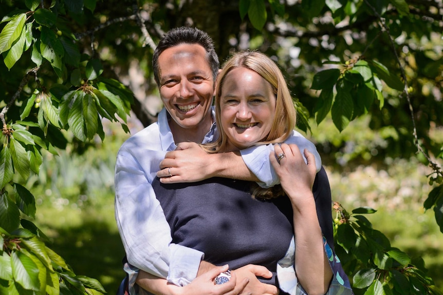 A man has his arms around a woman, both smiling broadly in front of a leafy background.