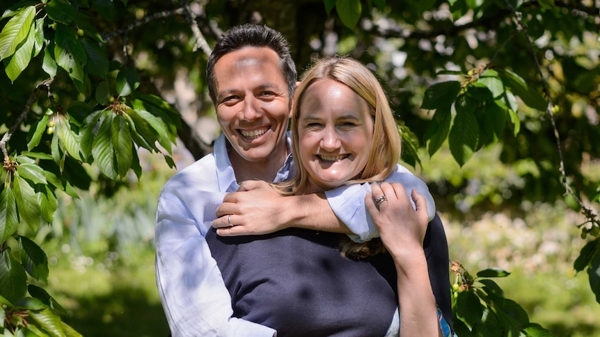 A man has his arms around a woman, both smiling broadly in front of a leafy background.