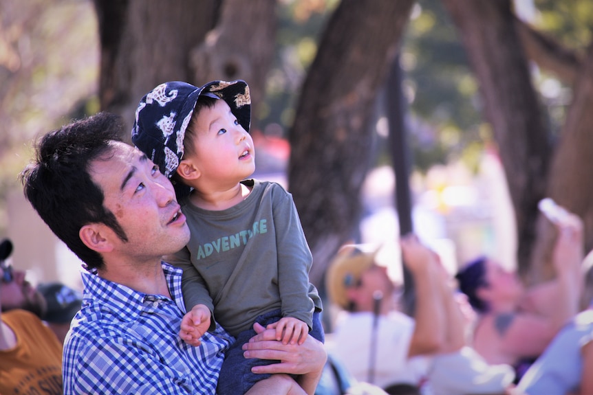 A man holds his son and looks up at the RAAF practice flyovers.
