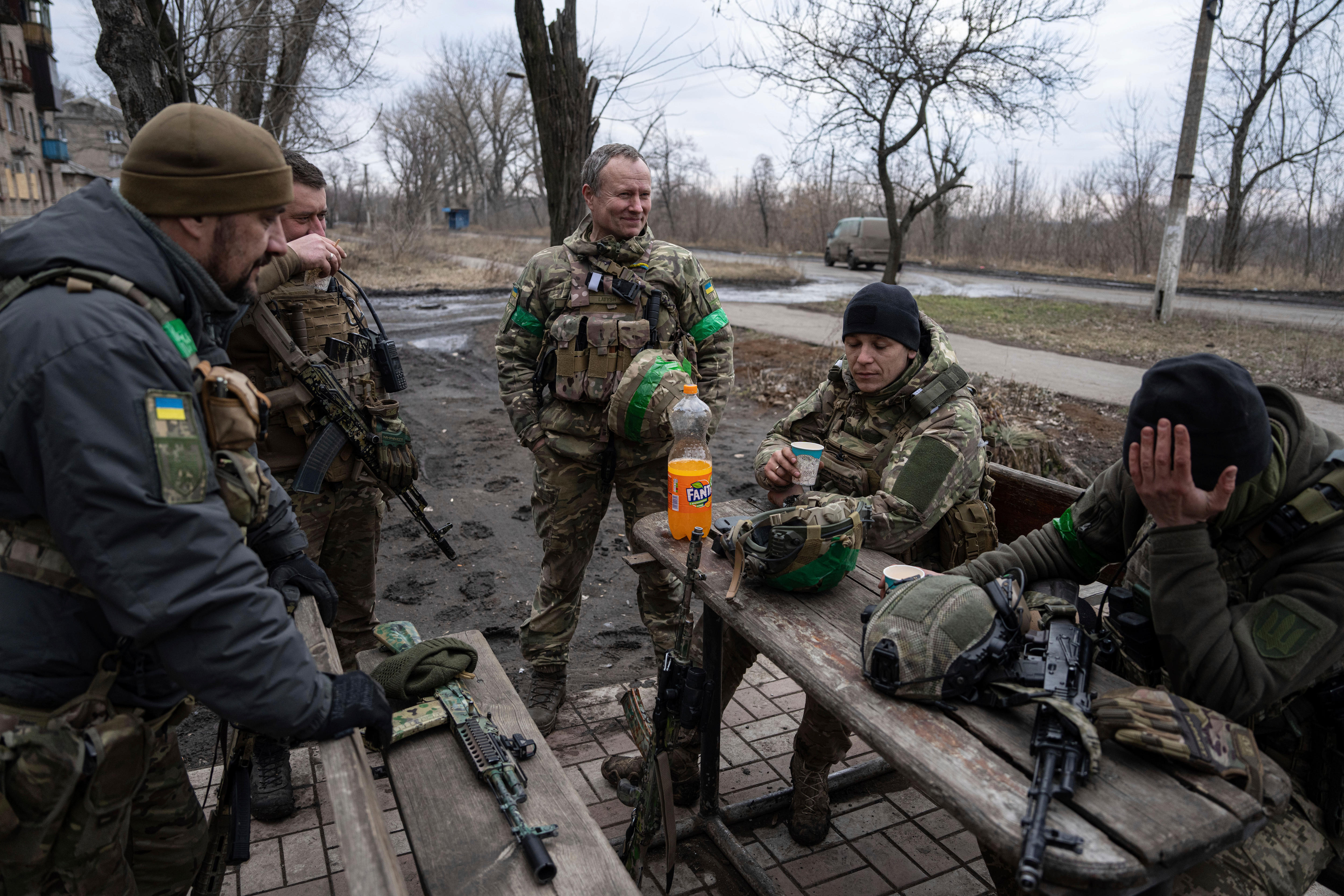 A group of men in combat gear, without their helmets, sit and stand around a park bench looking exhausted.