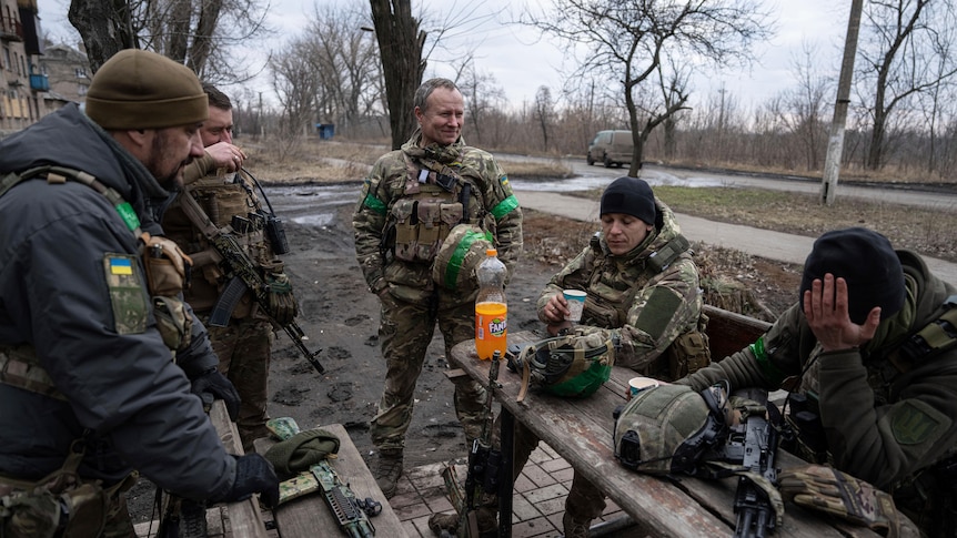 A group of men in combat gear, without their helmets, sit and stand around a park bench looking exhausted.