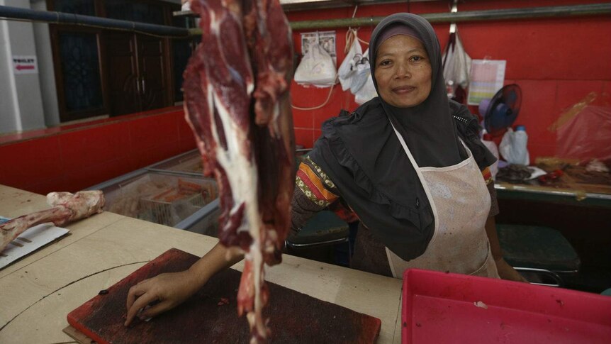 A woman in a headscarf sits behind a piece of hanging beef at a butcher's stall.