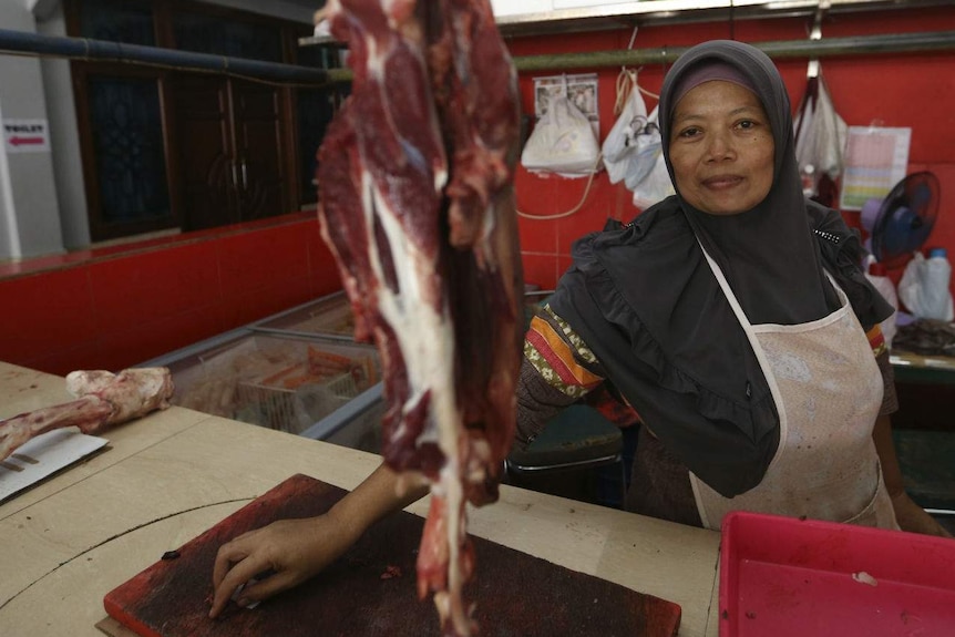 A woman in a headscarf sits behind a piece of hanging beef at a butcher's stall.