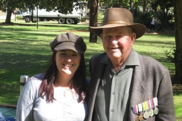Woman with long brown hair, grey cap and whit shirt next to an elderly man wearing a wide brim hat and jacket with war medlas
