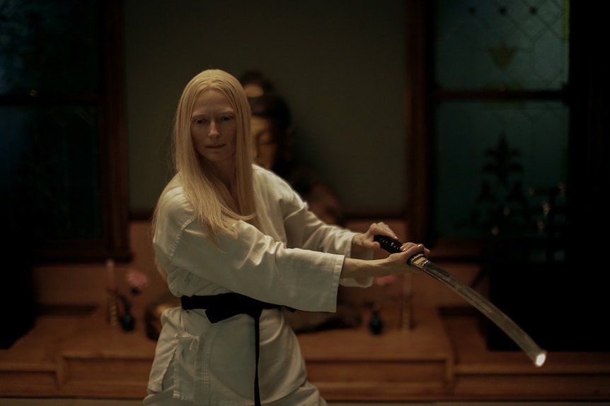 A woman with serious and focused expression wears all white martial arts attire wields a samurai sword in dimly lit room.