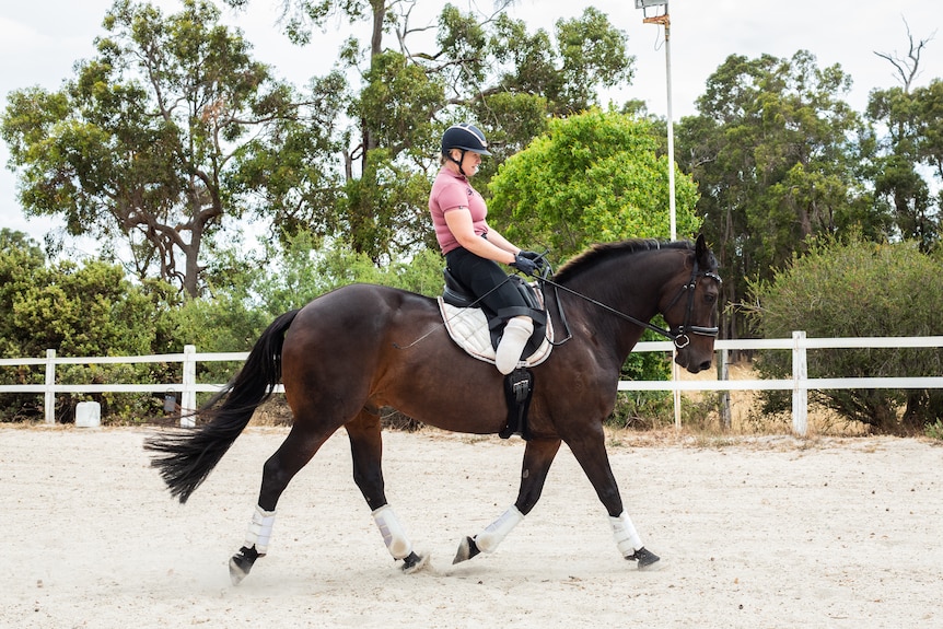 A woman with her leg amputated below the knee rides a black horse in a sand arena surrounded by trees