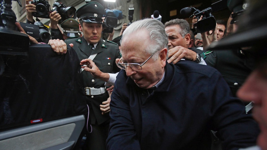 Fernando Karadima starts to bend to get into a car, as a uniformed man looks on, and the pair are surrounded by video cameras