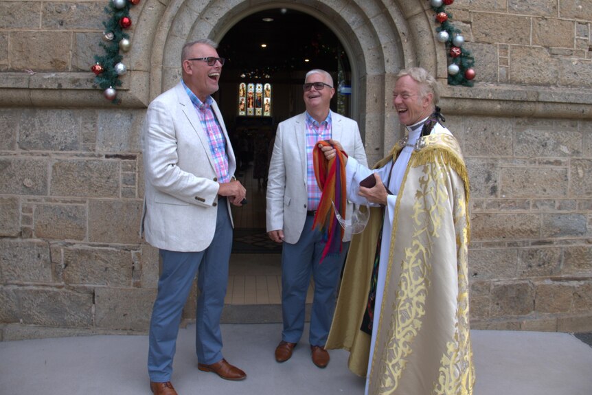 Two men in matching suits and a priest stand laughing outside a church. 